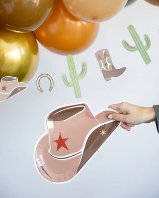 A hand holds a Cowboy Tan Hat Shaped Plate from My Mind’s Eye decorated with a red star. Balloons and other western-themed paper cutouts, including cactus and boot shapes, are in the background.