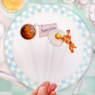 A hand holding three of Cami Monet's Diner Essentials Stir Sticks: one featuring a coffee cup, another with a Sweet'N Low packet, and the third adorned with an egg and bacon, all in front of a checkered plate.