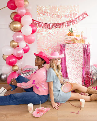 Two women in casual attire sitting on the floor at a pink-themed party with gold foil balloons and decorations, including My Mind's Eye Disco Ball Paper Plates.