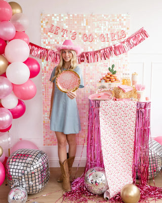 A woman in a party room decorated with "let's go girls" banner, pink balloons, My Mind’s Eye Disco Ball Paper Plates, disco balls, and a table with snacks and gold foil party plates, holding a pink tennis racket.