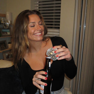 A woman in a black top smiles as she opens a beer bottle using the NPW Disco Bottle Opener, making it the perfect party gift.