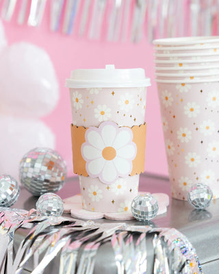 A pink My Mind’s Eye Disco Daisy To Go Cup with a daisy design and white lid stands among similar party cups and silver tinsel decorations. Small disco balls are also part of the display.