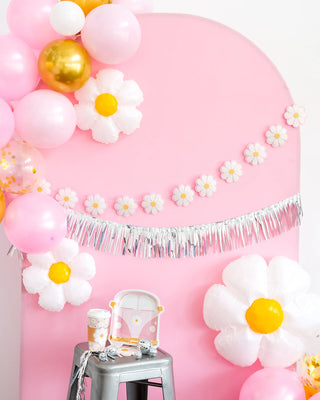 A pink backdrop adorned with pink, white, and gold balloons, white flower decorations, a silver fringe garland, and a small snack table with **My Mind’s Eye Disco Daisy Van Shaped Paper Plates** featuring whimsical designs sits next to a vintage van.