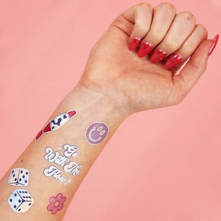 A hand with red nails displays NPW's Disco Temporary Tattoos on the forearm, featuring a rainbow, smiley face, "Go With The Flow" text, dice, and flower on a pink background.