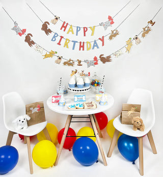 A dog-themed party setup with animal decorations, a colorful "Happy Birthday" banner, a dessert table featuring cupcakes, gift bags, and chairs, all surrounded by red, blue, and yellow balloons. The playful Good Dog Party Garland from Merrilulu completes the festive look.