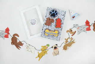 Dog-themed party decorations include the Merrilulu Good Dog Party Garland with cartoon dog cutouts, a framed display featuring art of paw prints, bones, and hydrants, and a small party hat for your pet.