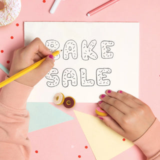 Close-up of hands writing "Bake Sale" in bubble letters on a sheet of paper, surrounded by craft supplies and Fred and Friends Donut Erasers with a vanilla scent on a pink background.