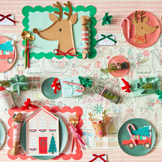 Festive table setting with reindeer and house-themed plates, wrapped presents, candy canes, and Christmas decorations in red, green, and gold hues. Meri Meri's Driving Reindeer Napkins add a lively touch to the holiday tableware.