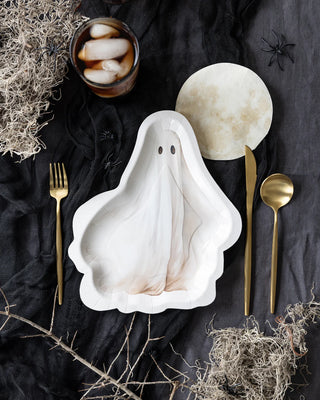 A My Mind’s Eye "Ghost Plate - Neutral" with a cute face sits on a dark tablecloth, perfect for any Halloween party. It is accompanied by a fork, knife, spoon, drink with ice, and dried plant decorations. A circular paper plate is also present, completing the spooky yet charming setup.