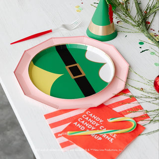 A holiday party table setting features an elf's outfit Christmas-themed paper plate, a plastic fork, a candy cane, a colorful party hat, and Bonjour Fête's Elf Candy Small Red Napkins with festive text. Greenery and confetti are scattered on the table for added cheer.