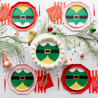 Top-down view of a holiday party table with elf-themed plates, a decorated cake featuring an elf belt design, Bonjour Fête's Elf Candy Small Red Napkins, candy canes, miniature elf hats, utensils, and festive greenery.