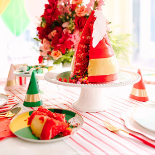 A red and yellow triangular cake on a white cake stand, with slices cut out revealing its layered interior, is surrounded by Bonjour Fête Elf Party Hats and a floral arrangement, creating a festive Christmas party vibe on the decorated table.