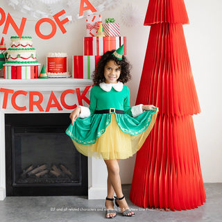 A child, adorned in an Elf Party Hat by Bonjour Fête and a green dress with a yellow skirt, stands beside a large red Christmas tree decoration. The background, themed for a festive Christmas party by Bonjour Fête, showcases holiday decor and presents.