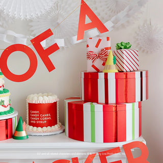 A display of wrapped gifts in red, white, and green, along with a decorated cake and candy, sets the scene for a Christmas party. Above the gifts and cake is a banner with red letters that reads "Bonjour Fête." Elf Party Hats from Bonjour Fête add a whimsical touch to the festive setup.