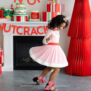 A girl in a pink Santa dress and candy cane shoes twirls joyfully at a Christmas party in a festive room adorned with a "Nutcracker" theme. Wrapped presents, Elf Party Hats from Bonjour Fête, and a toy elf on the mantle set the scene for this joyful celebration.