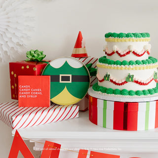 A festive holiday party display includes a tiered cake with green frosting, wrapped gifts, a decorative plate, a striped hat, and a red sign reading "CANDY, CANDY CANES, CANDY CORNS, AND SYRUP." Adding an extra touch of cheer and responsibility are eco-conscious materials and Bonjour Fête's Elf Candy Small Red Napkins.