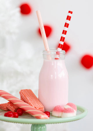 A glass bottle of pink milk with two Elf Reusable Straws by My Mind’s Eye, surrounded by red wafer biscuits, candy canes, red candy, and heart-shaped marshmallows on a green plate—perfect for Christmas celebrations.