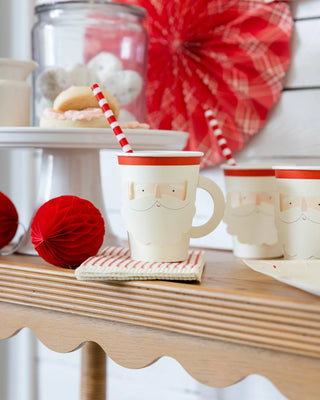 Festive holiday-themed table setting featuring paper cups with Santa faces, Elf Reusable Straws by My Mind’s Eye in red and white stripes, a cake stand brimming with treats, and red decorative orbs, all beautifully arranged on a wooden table—a perfect touch for Christmas celebrations.