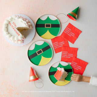 A child's hand reaches for a slice of cake on plates designed with elf costumes. Nearby, Elf Candy Small Red Napkins and hats labeled "Candy, Candy Canes, Candy Corns, and Syrup" adorn the festive holiday party table. The cake reads "SON NUTCRACKER" in icing. Bonjour Fête stands out as the ultimate party touch.