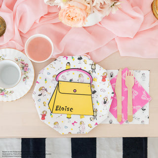 A table adorned with various objects including teacups, a pink napkin with utensils, a floral plate, and an Eloise Large Plate from Daydream Society, shaped like a yellow handbag emblazoned with the name "Eloise." Flowers and a pink cloth complete this charming setup, evoking the whimsical elegance one might find at the Plaza Hotel.