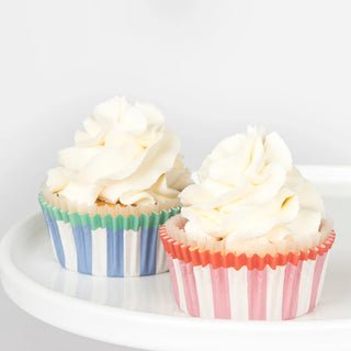 Two cupcakes, crafted by a champion cupcake maker, are topped with white frosting. One sits in a blue and green striped wrapper and the other in a pink and white striped wrapper, both from the Horse Cupcake Kit by Meri Meri. Delicate horse toppers adorn each cupcake on a white surface.