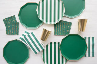 A collection of elegant party tableware made from eco-conscious materials, featuring Bonjour Fête Emerald Green Signature Cabana Stripe plates, solid green octagonal plates, napkins with decorative motifs, and gold cups, all neatly packed.