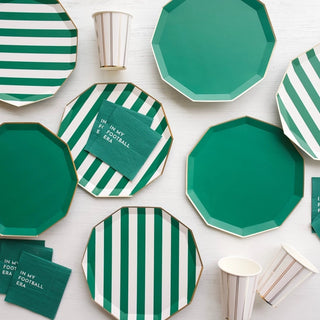 A collection of Bonjour Fête's Emerald Green Signature Cabana Stripe party tableware, including octagonal plates and paper cups made from eco-conscious materials, arranged on a white wooden surface, some featuring a football-themed phrase.