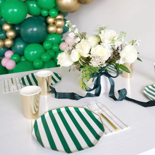 An elegantly set table featuring Bonjour Fête's emerald green signature cabana stripe plates, golden cups, cutlery, and a bouquet of white flowers, with a backdrop of green and pink balloons.