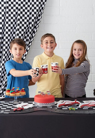 Three children holding Talking Tables F1 Red Race Car Paper Cups celebrate at a car-themed party with a red cake and decorated cupcakes on a checkered tablecloth.