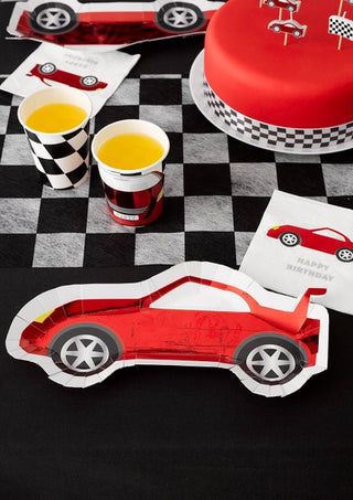 A race car-themed kids party setup featuring a red race car cake, yellow drinks in F1 Red Race Car Paper Cups by Talking Tables, and race car-shaped plates and napkins on a checkered tablecloth creates the perfect F1 themed party.