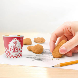 A hand uses a fried chicken-shaped eraser from Fred and Friends' "Fried Chicken Erasers" on a piece of paper, with a cup labeled "Fried Chicken Erasers" nearby, surrounded by scattered eraser pieces resembling chicken wings. An orange pencil rests alongside, ensuring your creativity stays BPA-free and TPR friendly.