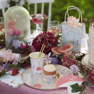 A beautifully decorated table is set for a fairy party with Meri Meri's Fairy Cups, floral-themed plates, and a cake. The table also features a glass cloche with flowers, pink tablecloth, golden foil stars, and various flora and whimsical fairy decorations.