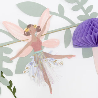 Create a magical atmosphere at your fairy party with the Meri Meri Fairy Garland, featuring illustrations of fairies with pink wings, pink tops, and flower crowns, all delicately suspended in mid-air amidst green leaves and purple honeycomb decorations.