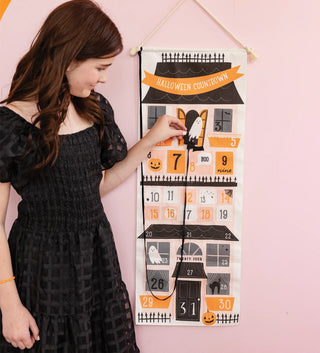 A girl in a black dress adjusts the numbers on the My Mind’s Eye Halloween Felt Countdown Advent calendar, adorned with decorative elements like ghosts, pumpkins, and the month of October prominently displayed.