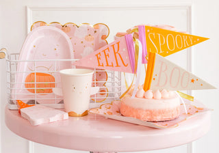 A small pink table with a decorated cake, Halloween-themed pennants, and My Mind’s Eye's Ghost With Basket Shaped Plates alongside paper cups and napkins featuring ghost and pumpkin designs, perfectly set up for a delightful Halloween party.