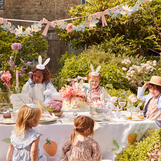 A group of children are sitting at a table in a garden surrounded by Meri Meri's Flower & Bow Garland.