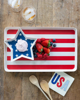 A patriotic-themed serving tray with a star-shaped bowl filled with whipped cream and berries, flanked by My Mind's Eye Foil USA Paper Cocktail Napkins, wooden spoons, and a glass of iced drink on a rustic wooden.