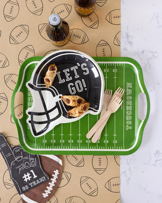 Set the scene for a tailgating party with a My Mind’s Eye Football Kraft Table Runner featuring a helmet-shaped plate, pastries, wooden cutlery, and a field-patterned tray. Completing the look are football-designed Kraft paper and beverage bottles in the background.