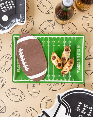 A top view of a table displays football-themed plates, a small football gift, two pizza slices, and a Football Kraft Table Runner by My Mind’s Eye, accentuated by decorated tablecloth and napkins.