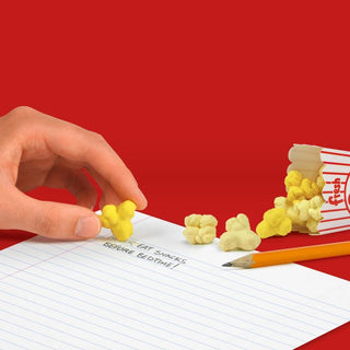 A hand is picking a piece of a buttery residue-free Fresh Popcorn Eraser from a red and white popcorn container on a piece of lined paper with a pencil. The paper has "Eat snacks before bedtime!" written on it, and the background is red. The product is from Fred and Friends.