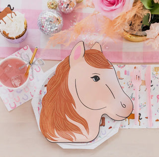 An image of a tabletop setting featuring a horse head-shaped paper plate, Daydream Society's Pony Tales Petite Napkins with a horseshoe pattern, a pink drink, a cupcake, and a floral arrangement in the corner.