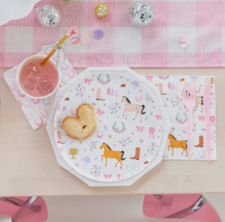 A table setting with Daydream Society's Pony Tales Small Plates, horse-themed napkins, and a placemat. There's a pink drink with ice, a heart-shaped toast, a pink fork and knife, small disco ball decorations on a pink and white tablecloth—perfect for your next pastel pony party.