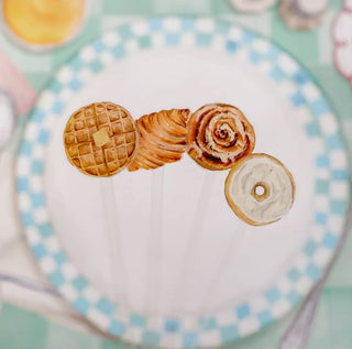 Four charming pastries on sticks—a waffle, croissant, cinnamon roll, and donut—reminiscent of a watercolor bouquet. Displayed on a white plate with a blue checkered rim, they capture the essence of Cami Monet's Breakfast Carbs Stir Sticks.