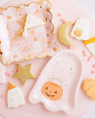 A pink table is set with a Ghost Scatter Plate from My Mind’s Eye, Halloween-themed pumpkin plates, ghost-shaped cookies, a star-shaped cookie, a moon-shaped cookie, and a triangular "BOO" cookie. Small pink candies are scattered around, creating the perfect spooky ambiance for your Halloween party.