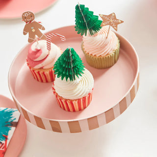 A charming display from the Gingerbread Cupcake Kit by Meri Meri features three cupcakes perched on a pink striped stand, complete with festive toppers such as a gingerbread man, candy cane, star, and paper Christmas trees.