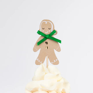 A gingerbread man-shaped paper decoration from the Meri Meri Gingerbread Cupcake Kit, adorned with a green bow and set on a swirl of white icing, makes an excellent festive topper. This charming gift enhances any holiday dessert display perfectly.