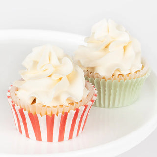 Two cupcakes from the Meri Meri Gingerbread Cupcake Kit, topped with whipped cream frosting—one nestled in a red-striped wrapper and the other in a plain green wrapper—rest on a white plate, embellished with festive toppers.