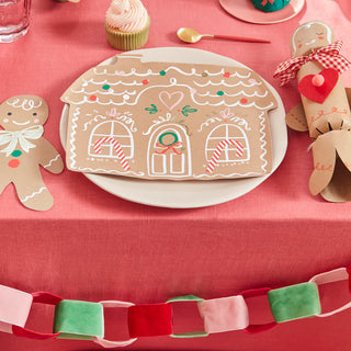 The festive table is set with Meri Meri's Gingerbread House Plates, two gingerbread man decorations, and a cupcake, all accented by a red-pink-green paper chain on the pink tablecloth. Perfect for a delightful Christmas party!