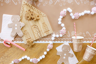 A festive setup featuring a wooden gingerbread house, gingerbread cookies, a candy cane, and string decorations sits on a wooden surface. Two paper cups with holiday beverage accessories and My Mind’s Eye Gingerbread Reusable Straws complete the scene.