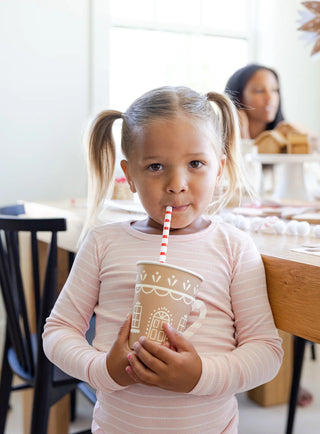 A young girl with pigtails sips from a patterned cup using a Gingerbread Reusable Straw by My Mind's Eye. Another person is blurred in the background, sitting at a table.